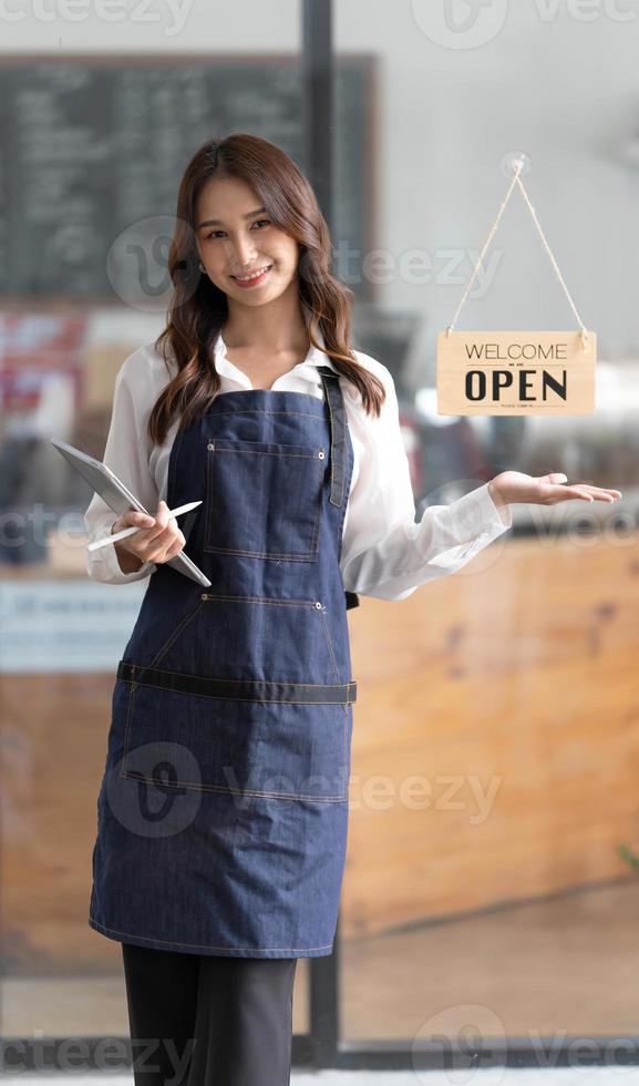 hermosa joven barista asiática en delantal sosteniendo una tableta y parada frente a la puerta de la cafetería con un letrero abierto. concepto de inicio de propietario de negocio. foto