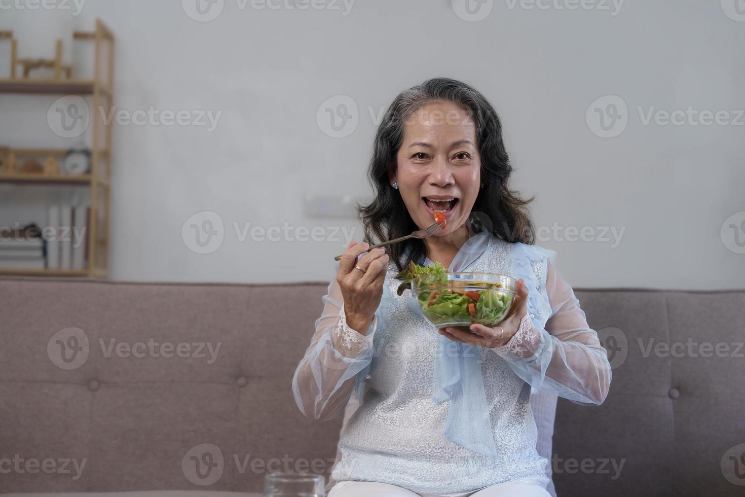 retrato, alegre mujer asiática de los años 60 desayunando en su sala de estar, comiendo un tazón saludable de ensalada de verduras. foto