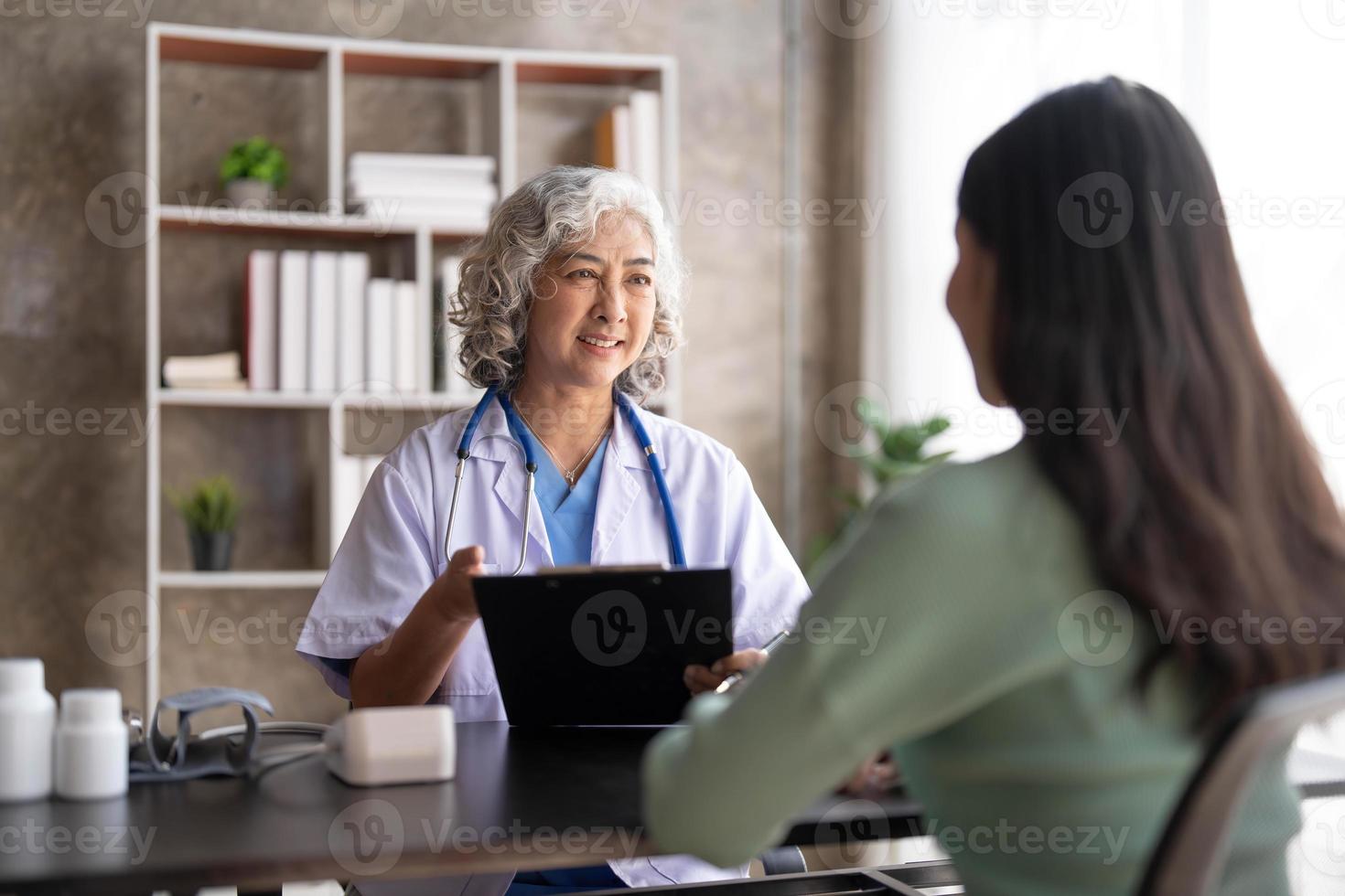 una doctora principal está leyendo el historial médico de una paciente y hablando con ella durante la consulta en una clínica de salud. médico en bata de laboratorio sentado detrás de una computadora portátil en la oficina del hospital. foto