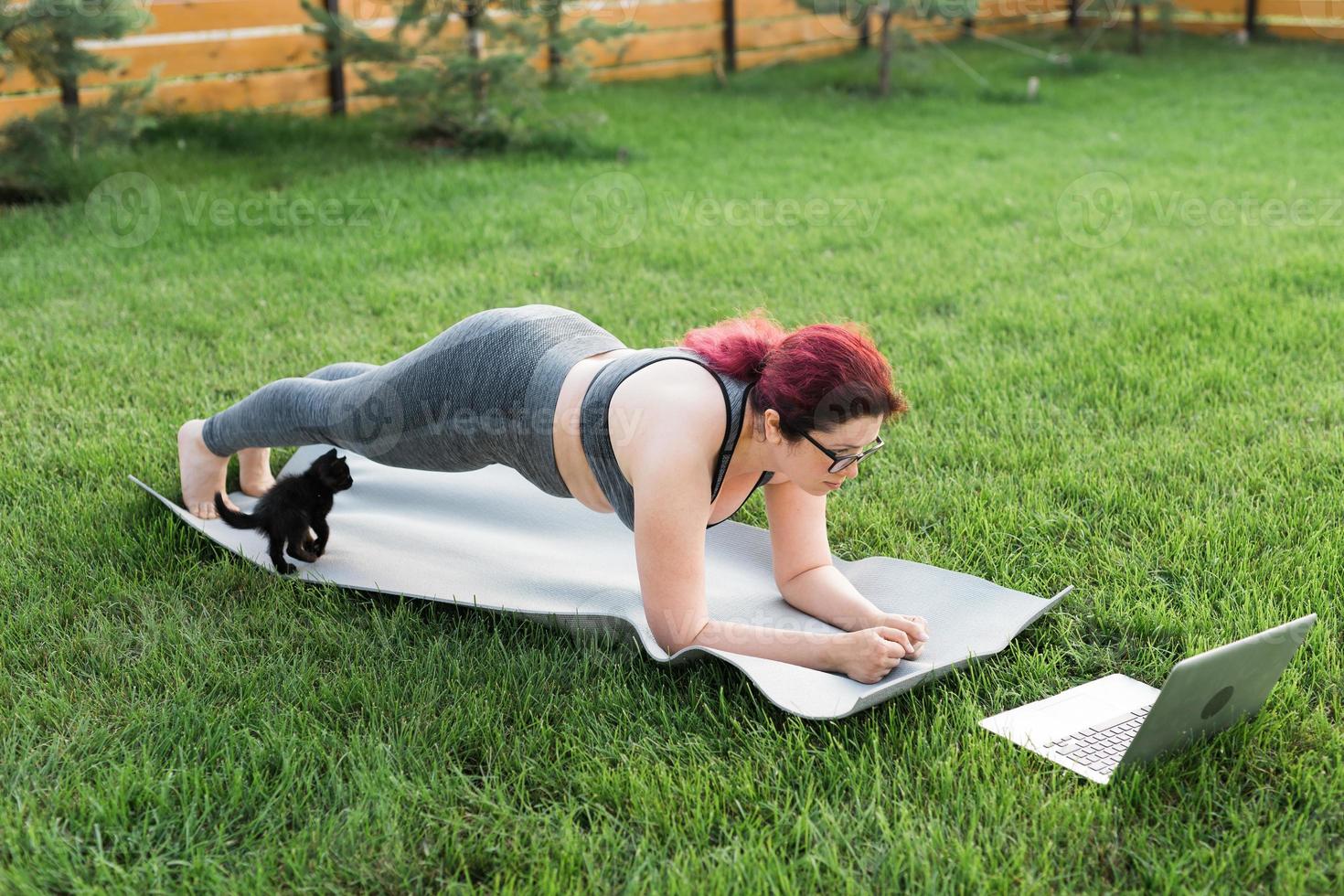 mujer joven de talla grande con top deportivo y polainas de pie en un tablón sobre una alfombra de yoga pasando tiempo en la hierba verde en el patio. gatito negro camina a su alrededor. concepto de bienestar y fitness foto