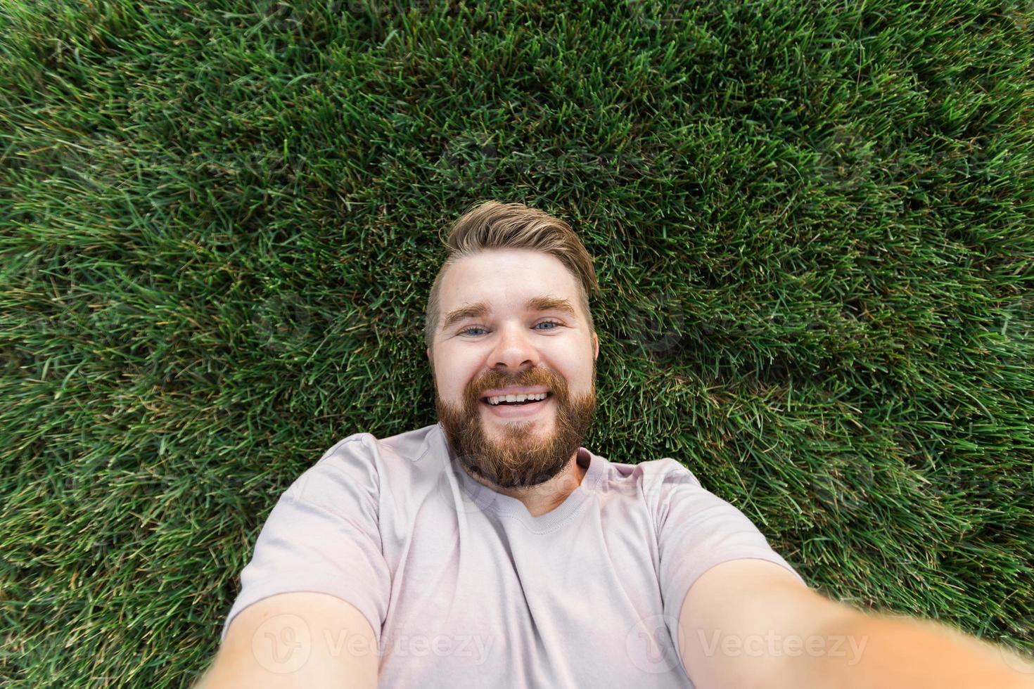 joven con barba y peinado de moda tirado en la hierba tomando selfie - sosteniendo un teléfono inteligente o una tableta y mirando la cámara. foto