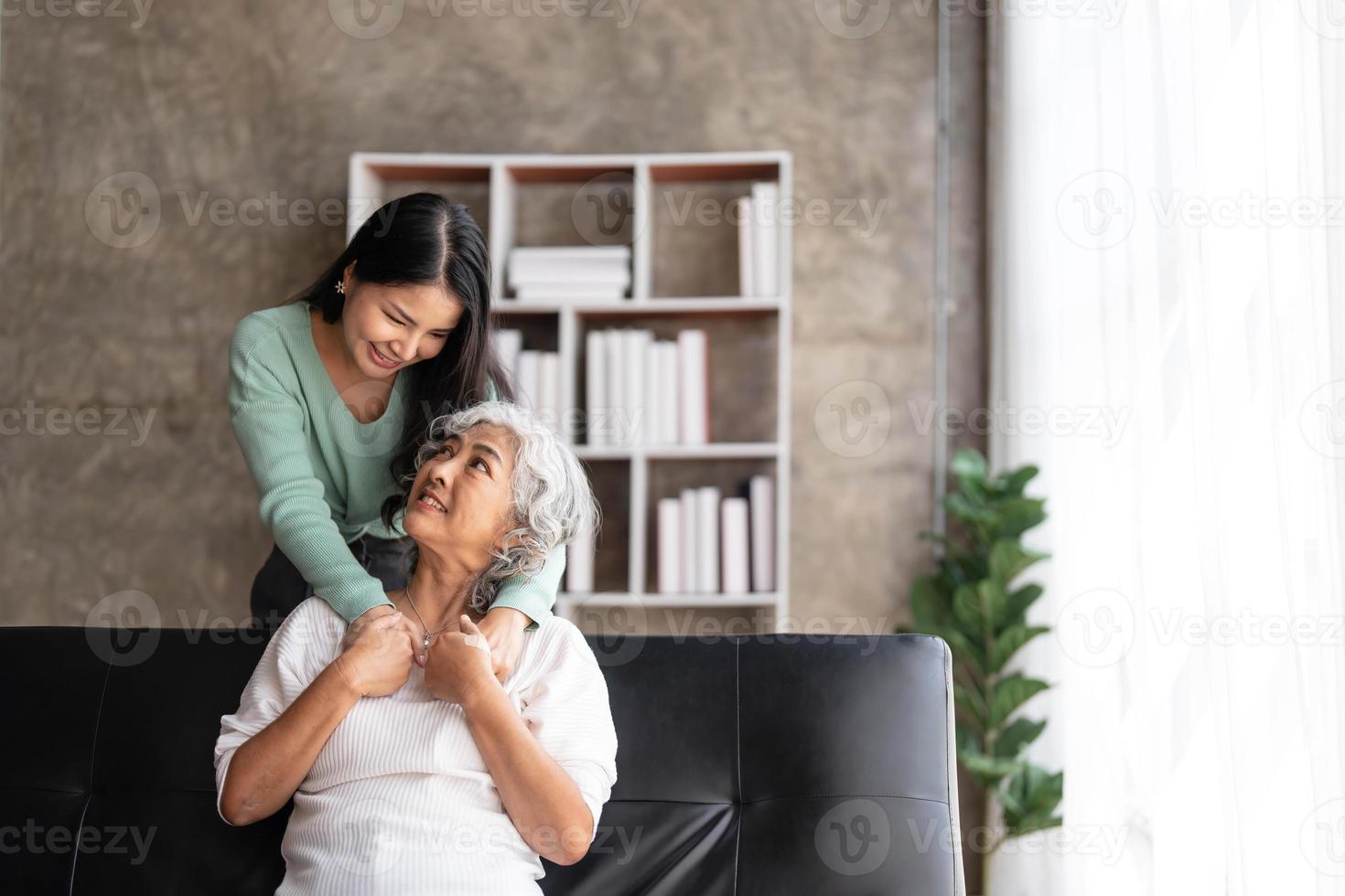 Loving adult daughter hugging older mother, standing behind couch at home, family enjoying tender moment together, young woman and mature mum or grandmother looking at each other, two generations photo