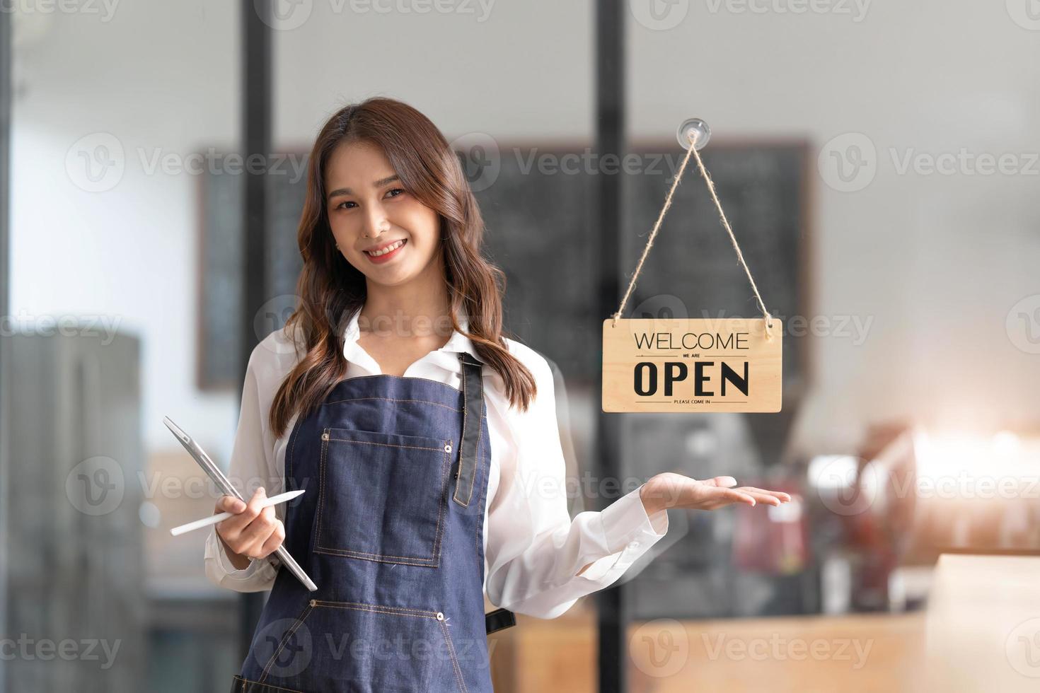 Beautiful asian young barista woman in apron holding tablet and standing in front of the door of cafe with open sign board. Business owner startup concept. photo