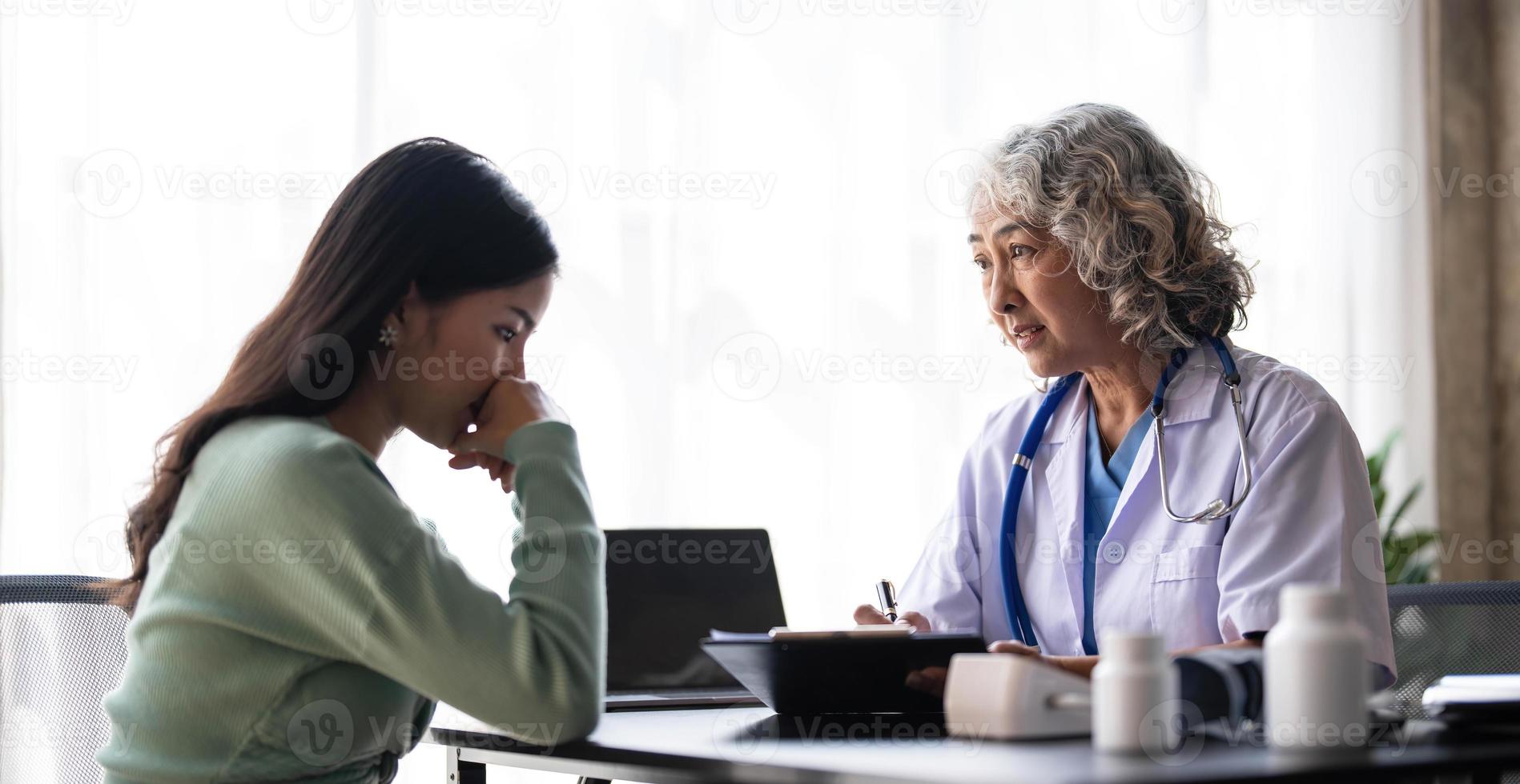 Woman senior doctor is Reading Medical History of Female Patient and Speaking with Her During Consultation in a Health Clinic. Physician in Lab Coat Sitting Behind a Laptop in Hospital Office. photo