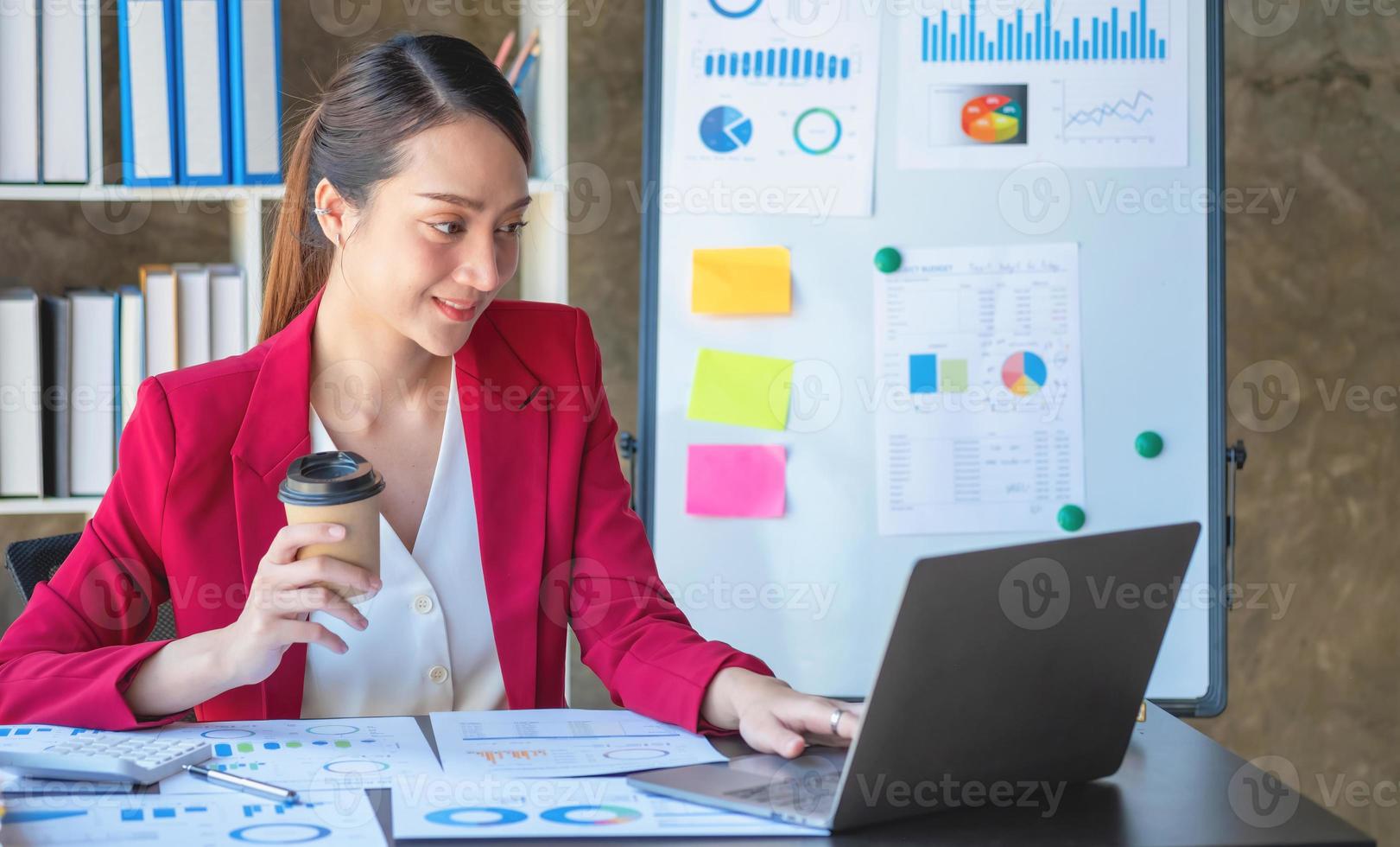 financiera, mujer de negocios asiática con traje rojo sosteniendo una taza de café sentada en el escritorio de la oficina, teniendo una computadora para hacer trabajo de contabilidad en el lugar de trabajo para calcular el beneficio anual por deber, idea de negocio foto