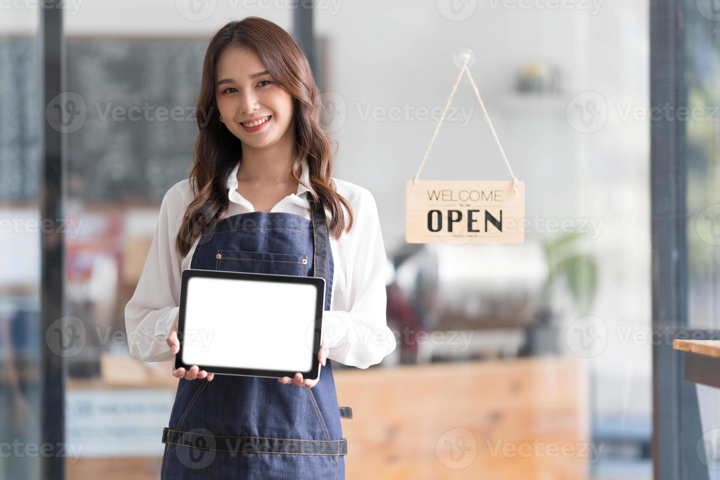 Beautiful asian young barista woman in apron holding tablet and standing in front of the door of cafe with open sign board. Business owner startup concept. photo