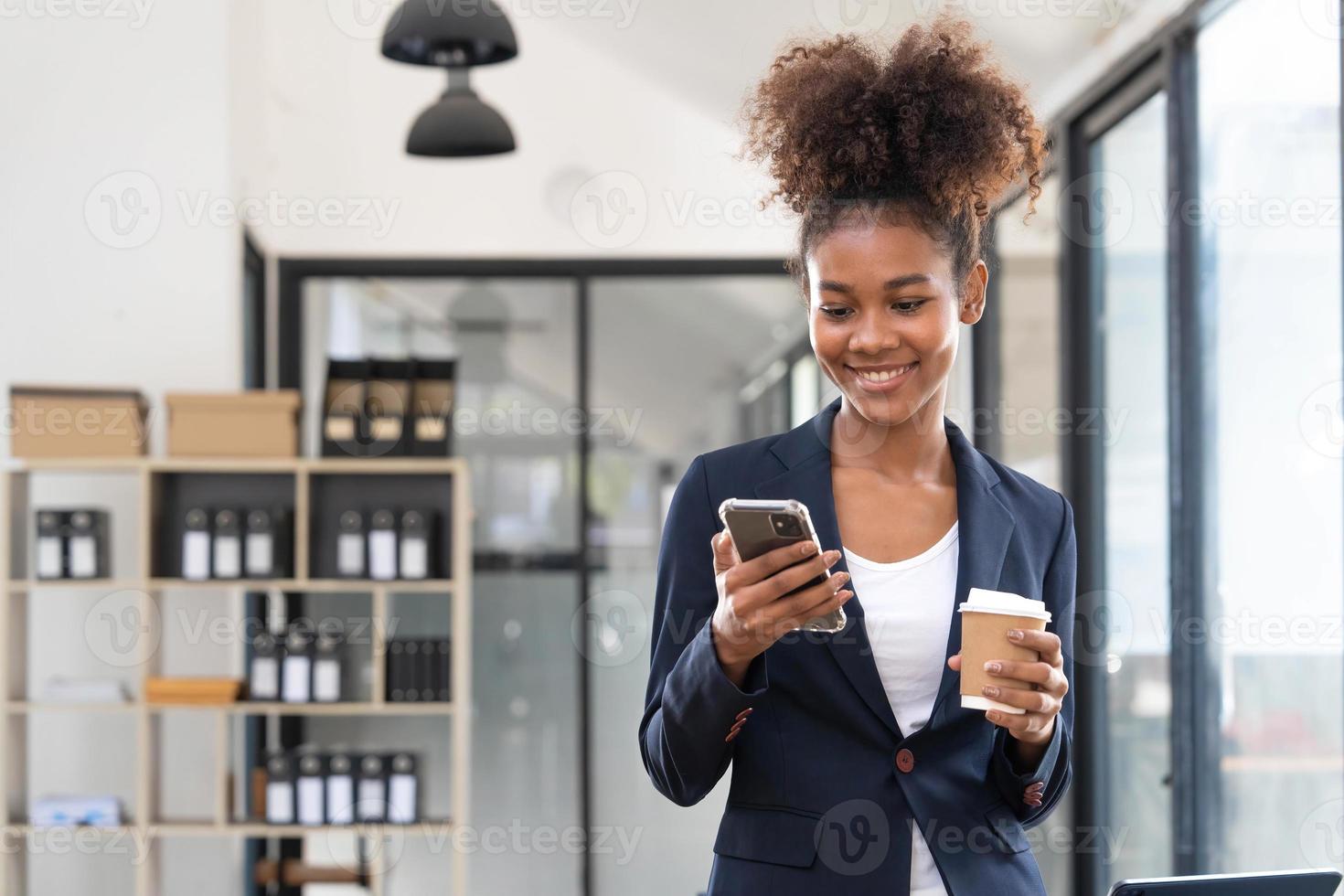 Shot of smiling young business woman sending messages with mobile phone sitting on the desk in the office. photo
