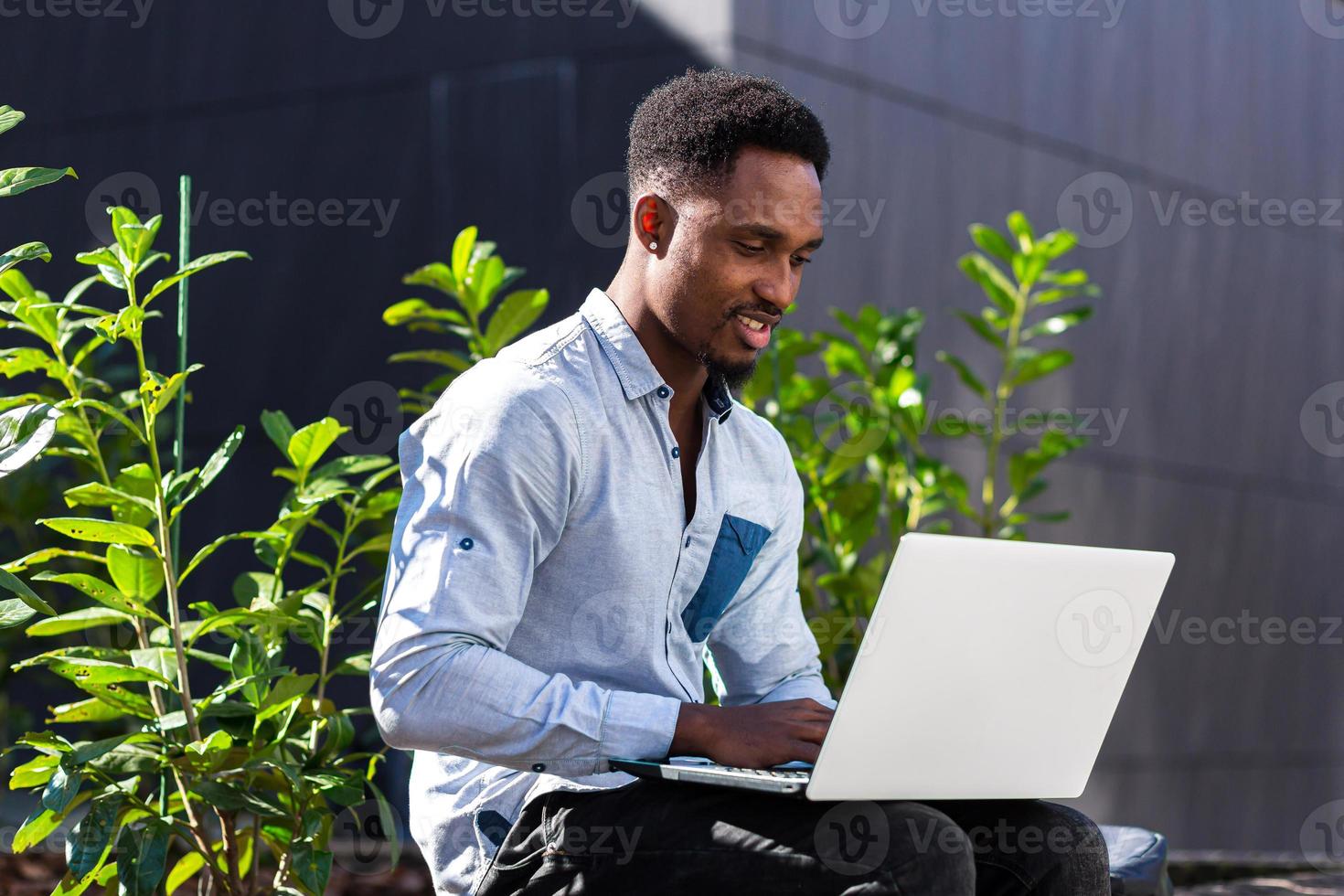 hombre negro independiente que trabaja en línea usando una computadora portátil sentada en un banco fuera de un edificio moderno de oficinas foto