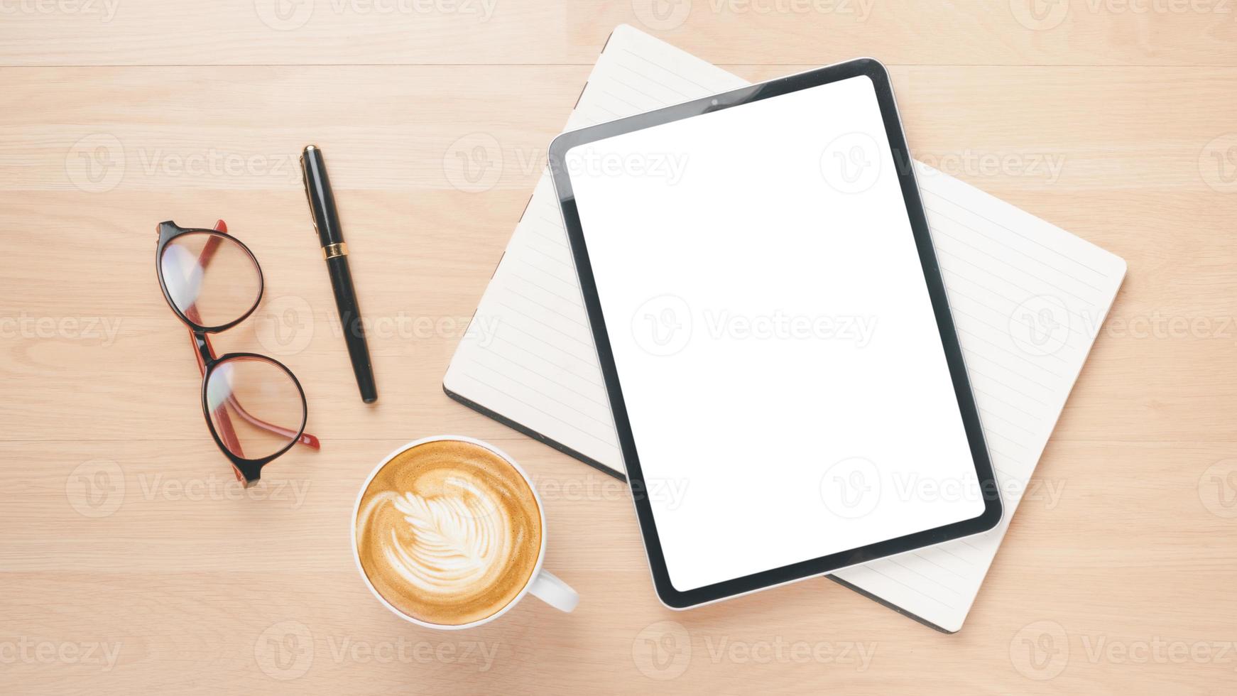 lugar de trabajo de escritorio de madera de oficina con tableta de pantalla en blanco, cuaderno, bolígrafo, anteojos y taza de café, vista superior plana. foto