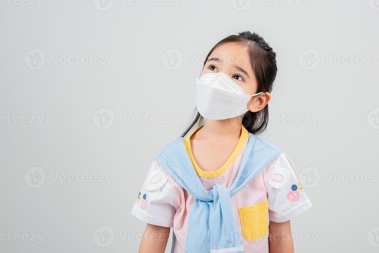 niña asiática con mascarilla respiratoria para proteger el brote de coronavirus y apuntando con la mano al fondo en blanco, nuevo virus covid-19 foto