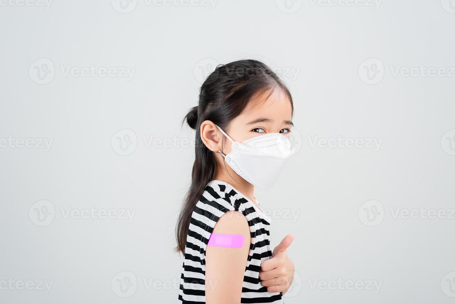 Asian little girl with medical mask showing her arm with bandage after virus vaccine. Kid and child vaccination campaign. Protect your kid with vaccine concept photo