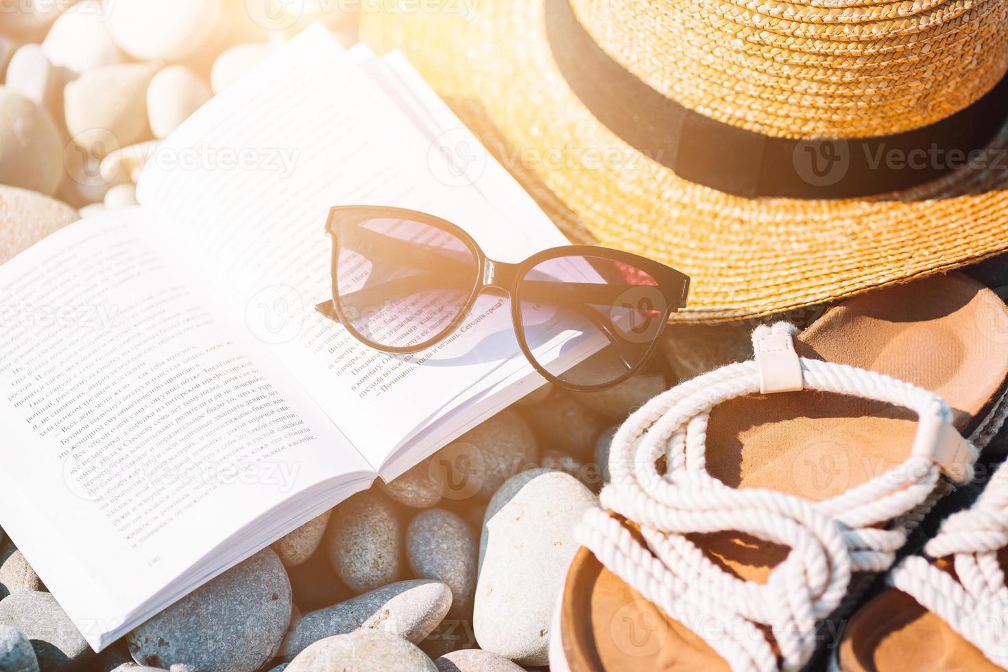 Beach hat on opened book with sunscreen and shoes on pebble beach photo