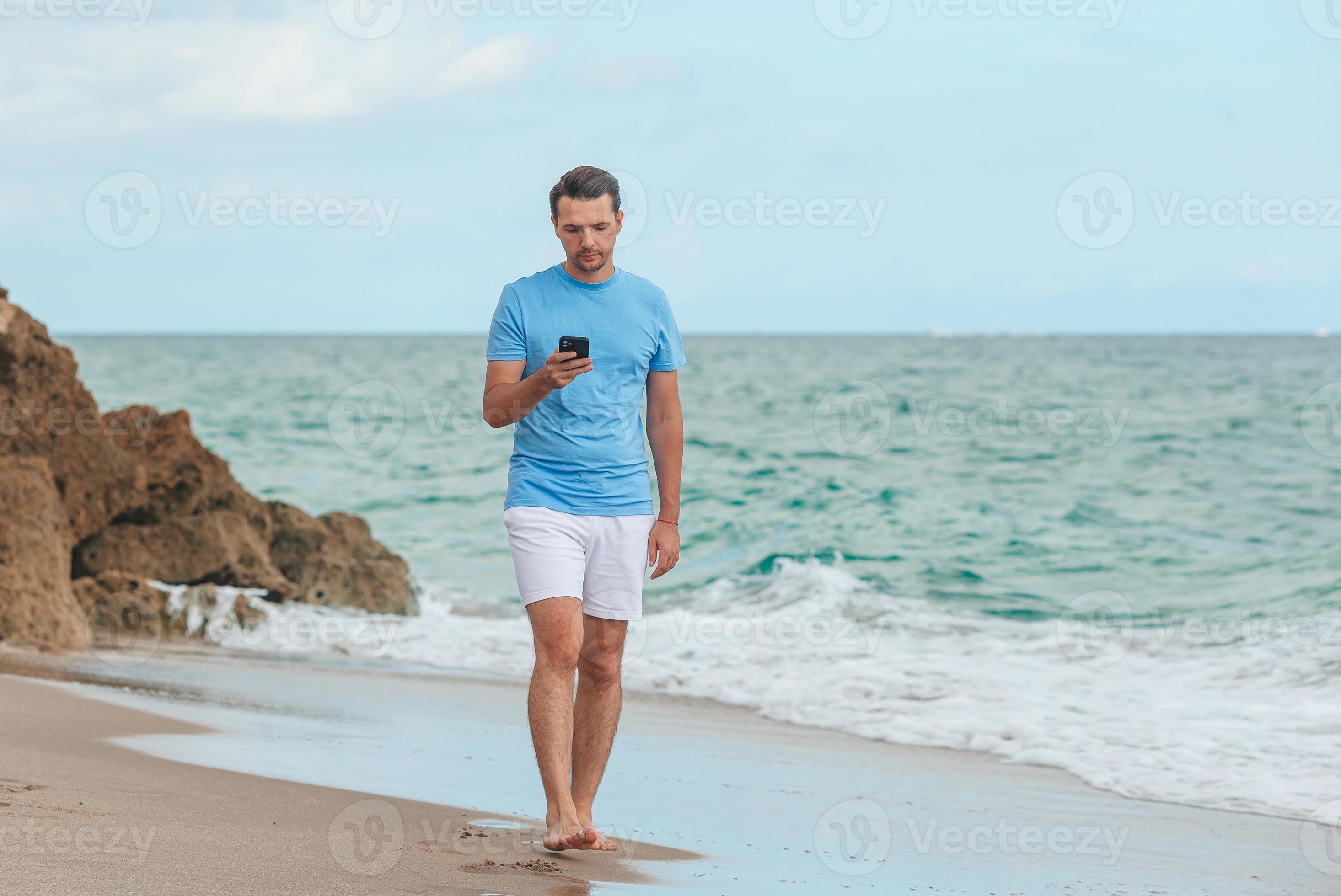 Young man holding mobile phone in his hand while walking on the beach