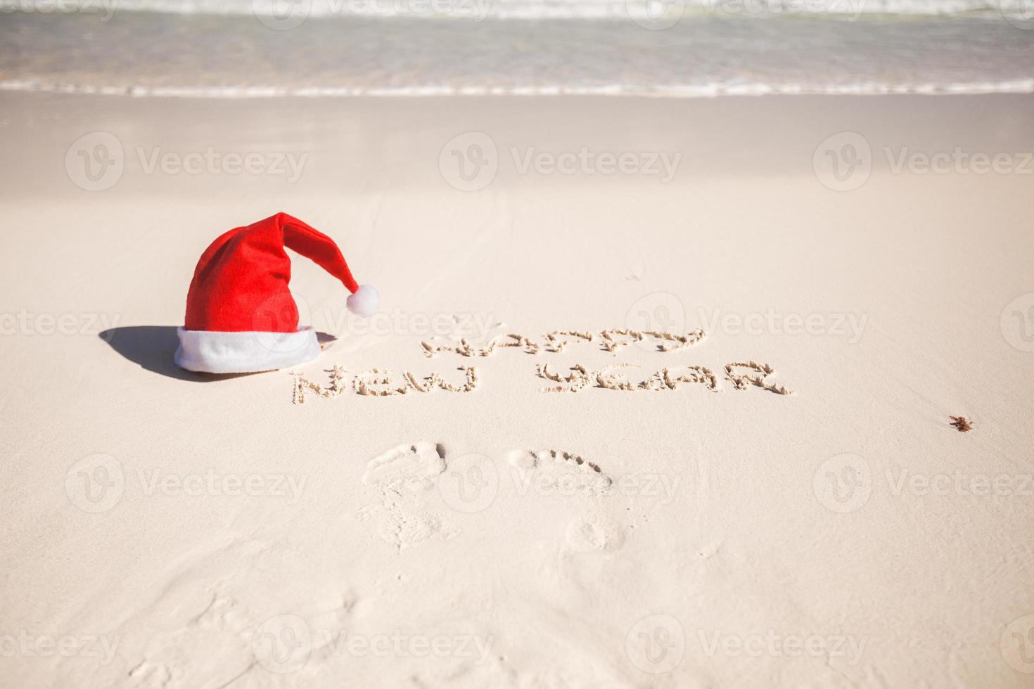 Happy New Year written in the sand and Santa Hat on white sandy beach photo