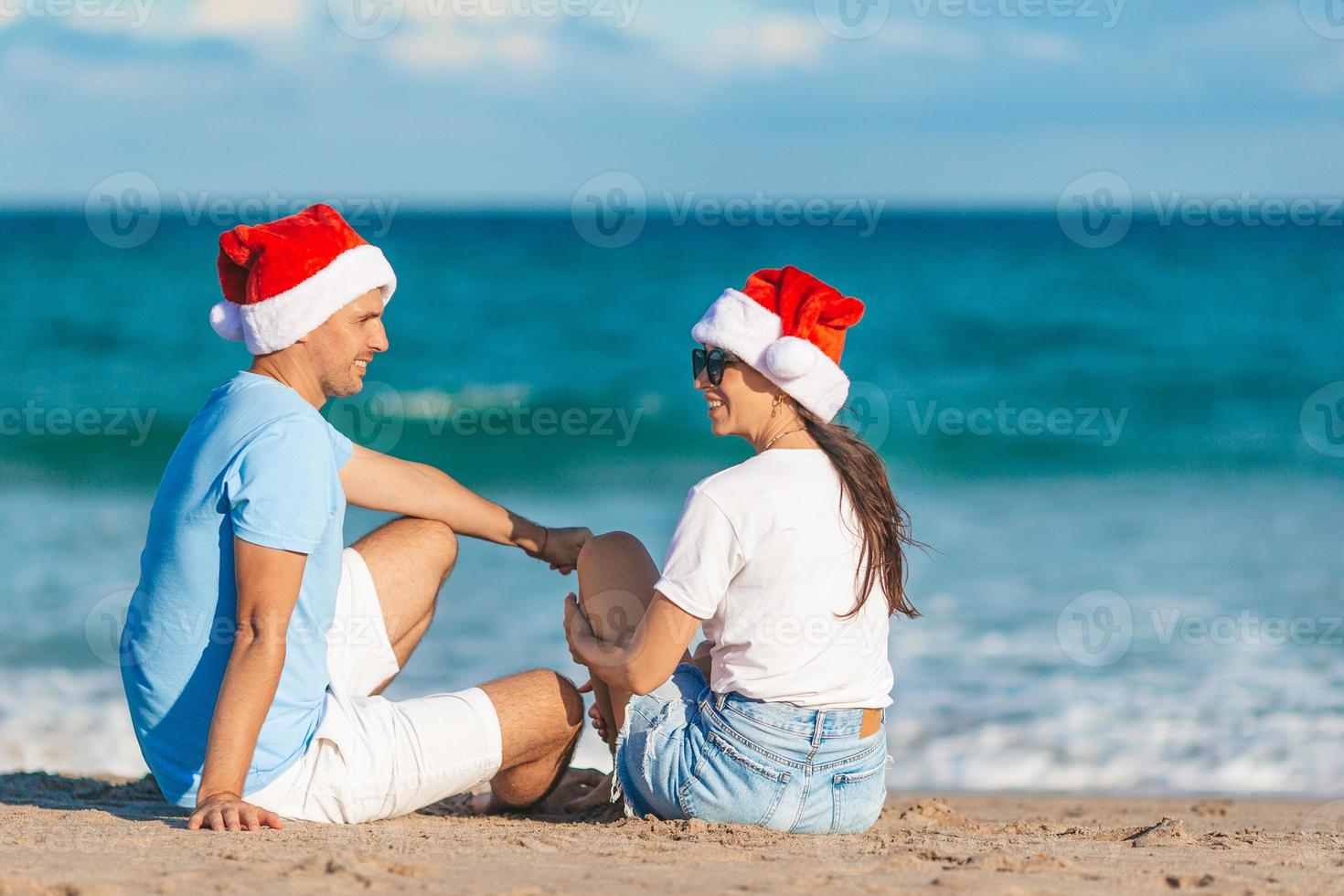 Young romantic couple in red Santa hats sitting on tropical white sand beach celebrating Christmas photo