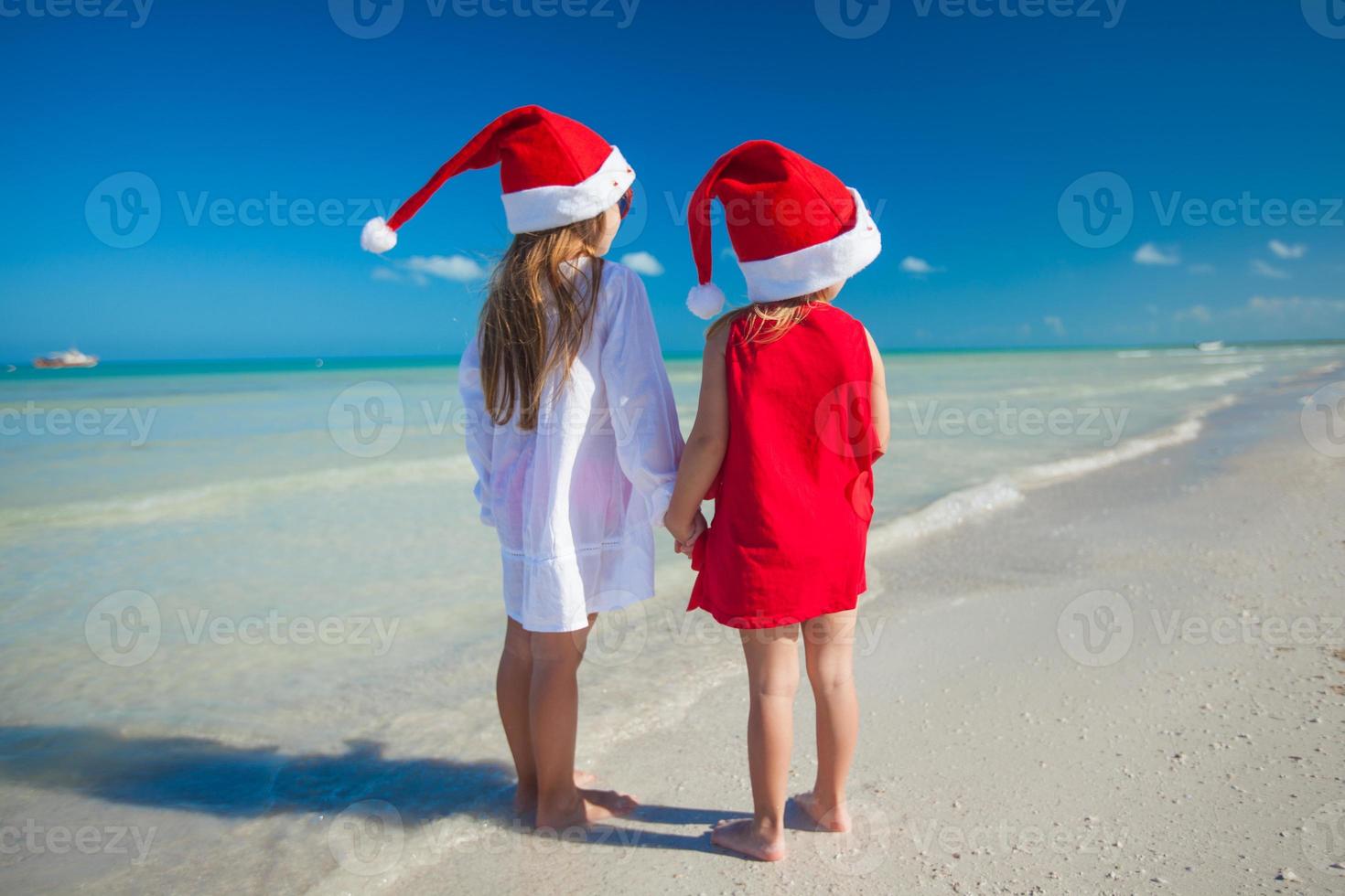 vista trasera de niñas lindas con sombreros de navidad en la playa exótica foto