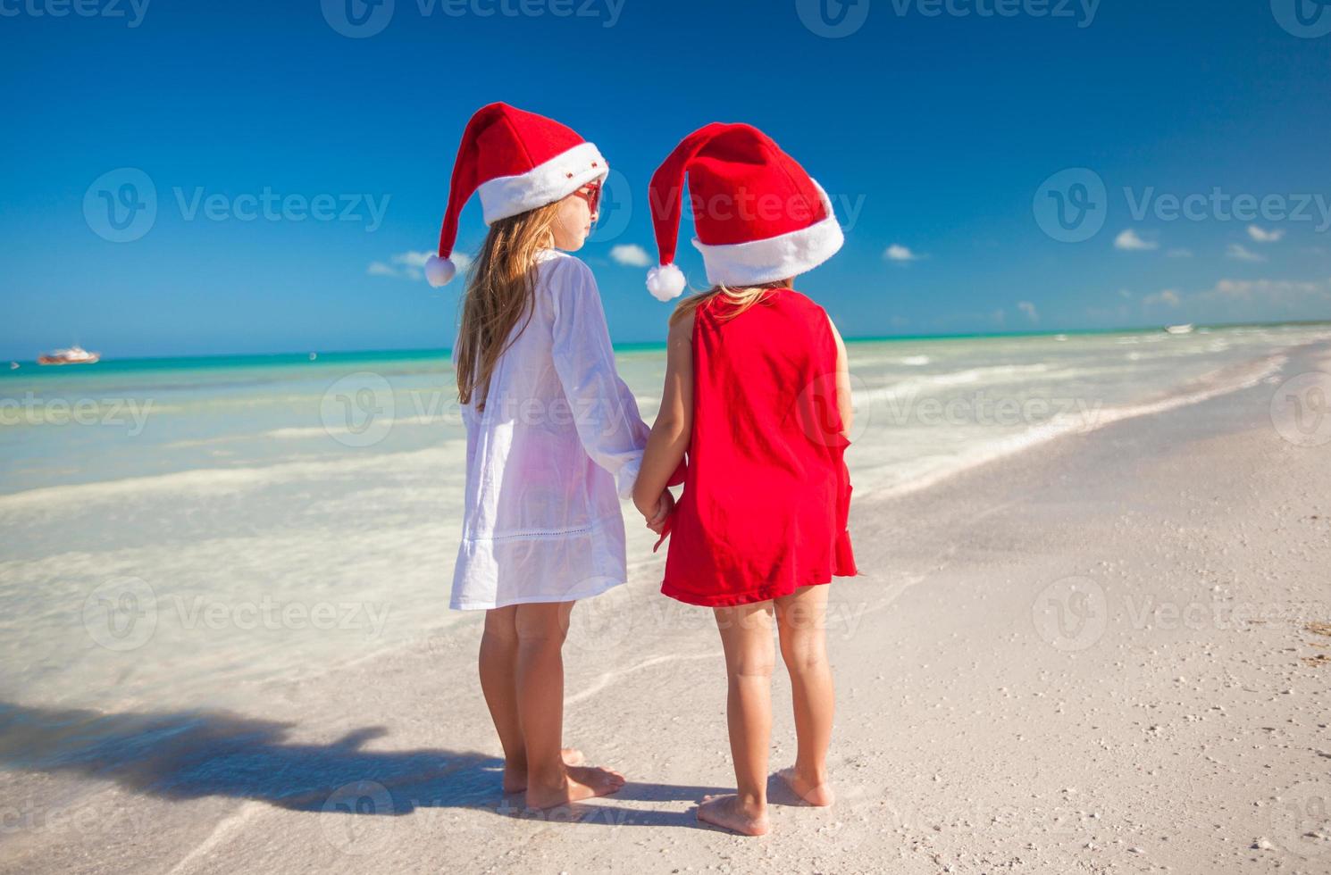 vista trasera de niñas lindas con sombreros de navidad en la playa exótica foto