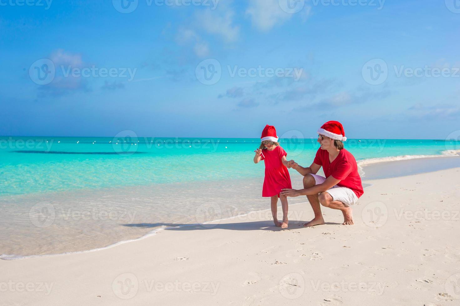 padre joven y adorable hijita con sombrero de navidad en la playa tropical foto