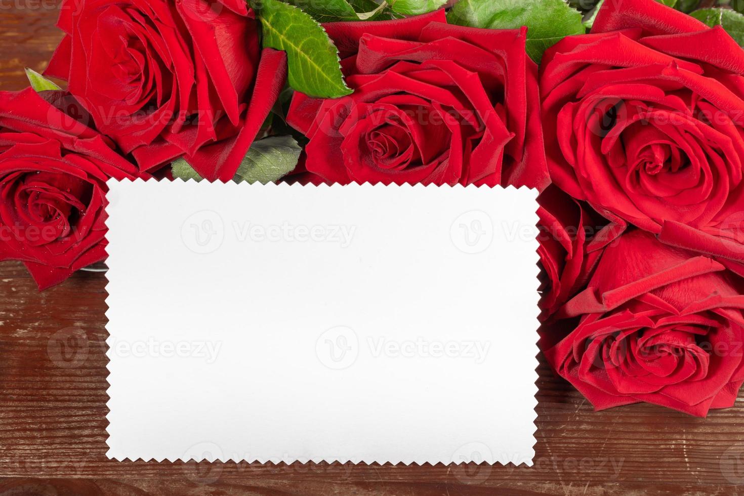 Red roses bouquet and white paper sheet for text on old rustic wooden table close up. Copy space. photo