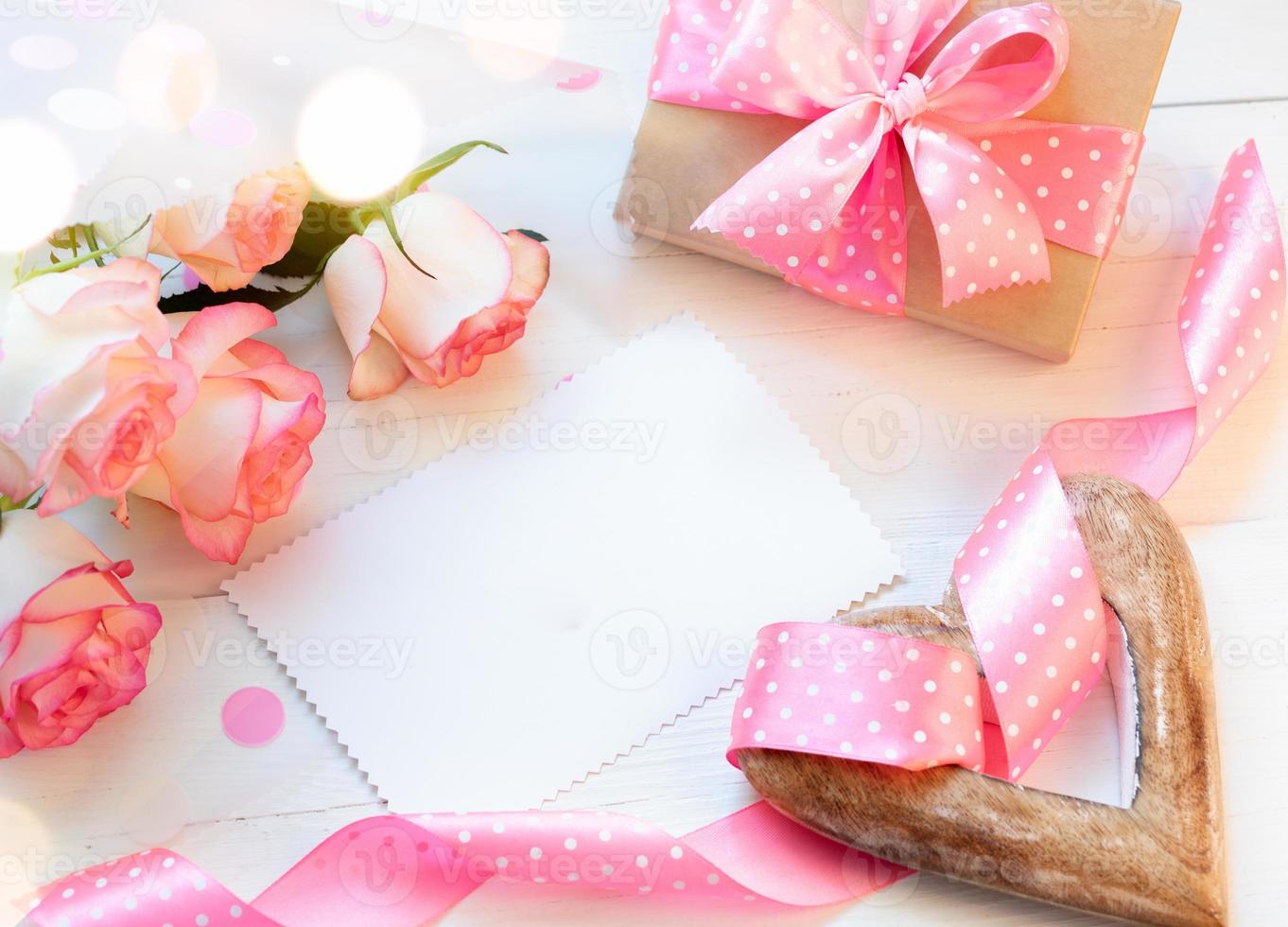 Frame of pink roses, gift box, wooden heart, pink ribbon polka dots, blank empty sheet on white. photo