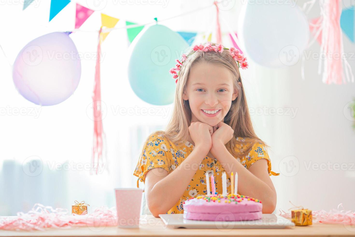 Caucasian girl is dreamily smiling and looking at birthday rainbow cake. Festive colorful background with balloons. Birthday party and wishes concept. photo