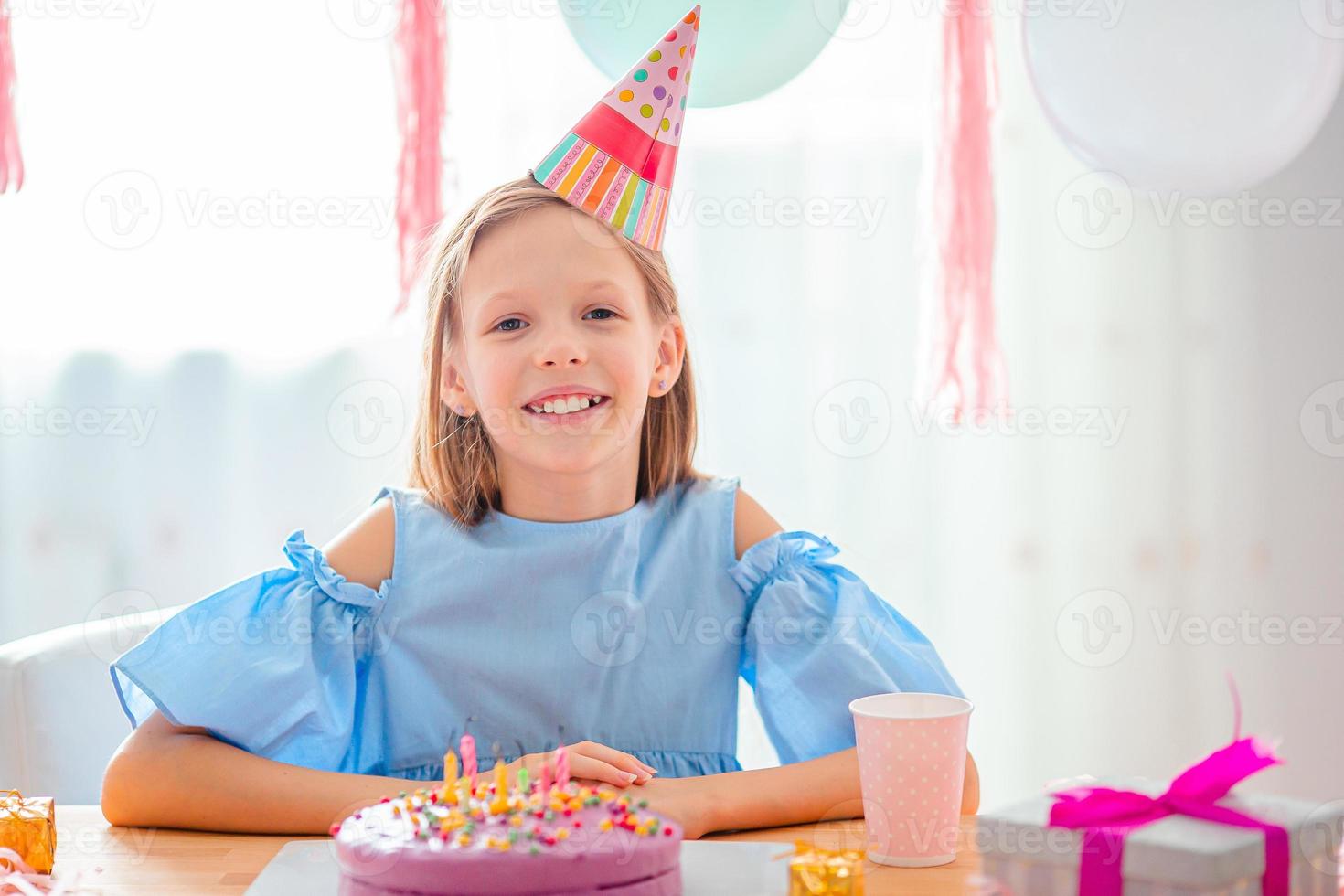 Caucasian girl is dreamily smiling and looking at birthday rainbow cake. Festive colorful background with balloons. Birthday party and wishes concept. photo