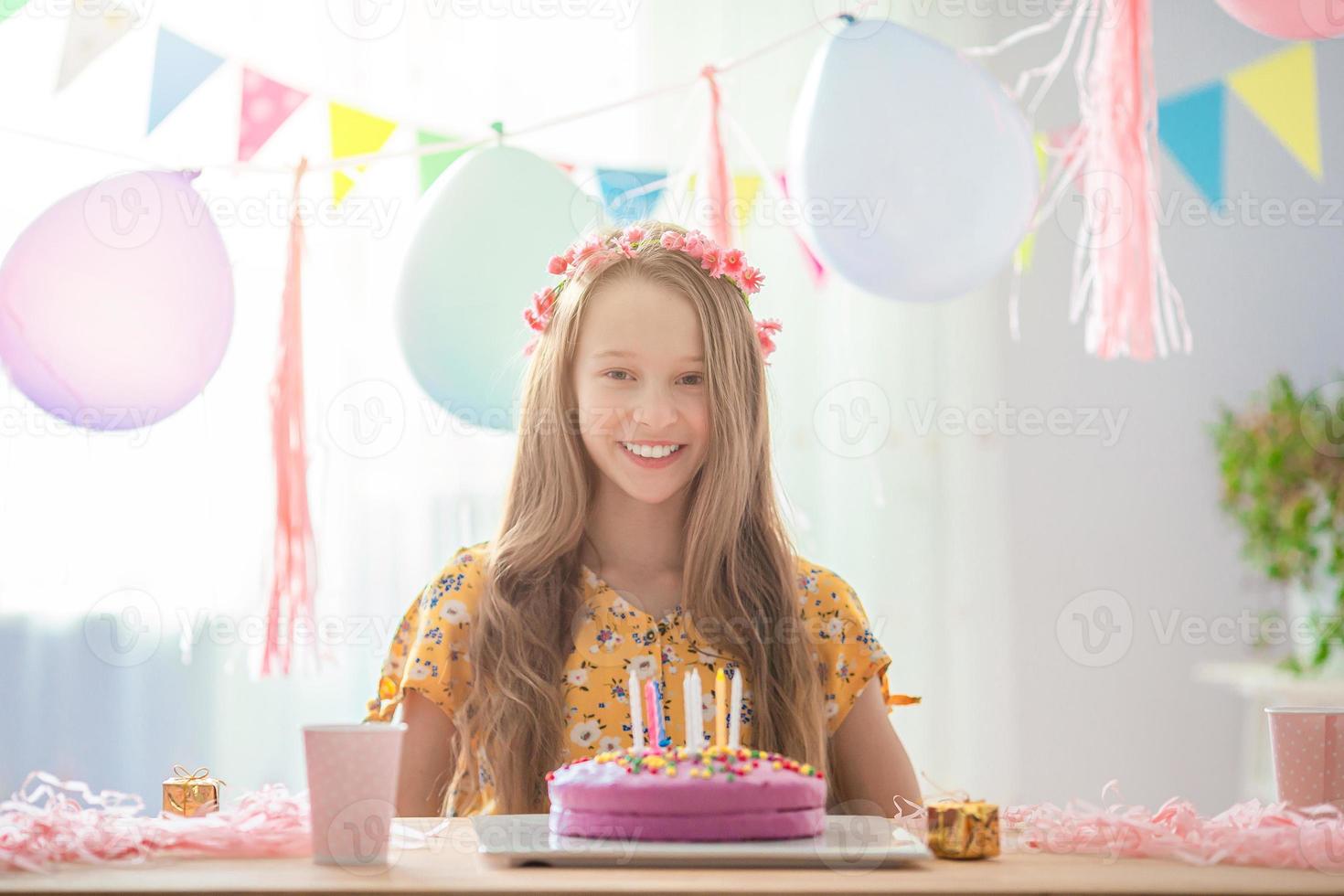 Caucasian girl is dreamily smiling and looking at birthday rainbow cake. Festive colorful background with balloons. Birthday party and wishes concept. photo