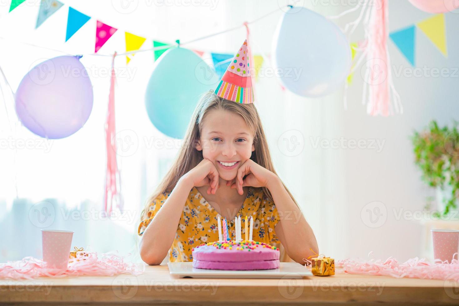Caucasian girl is dreamily smiling and looking at birthday rainbow cake. Festive colorful background with balloons. Birthday party and wishes concept. photo