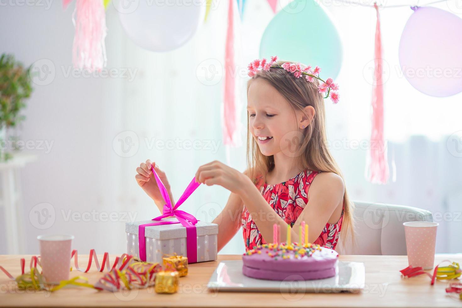 Caucasian girl is dreamily smiling and looking at birthday rainbow cake. Festive colorful background with balloons. Birthday party and wishes concept. photo