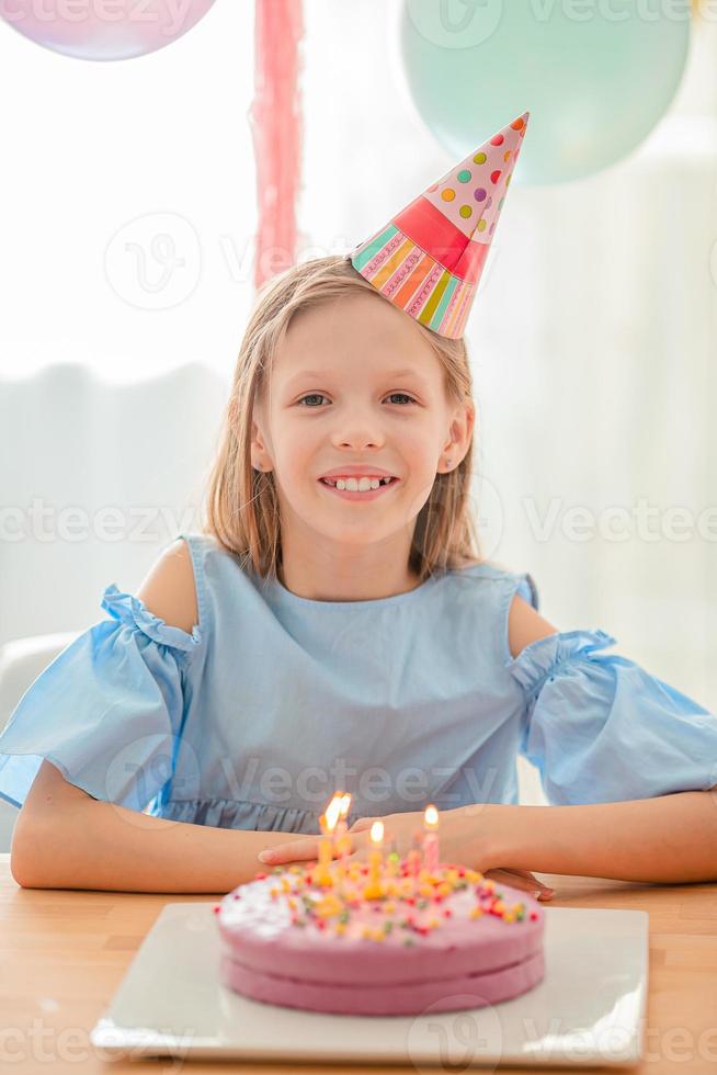 Caucasian girl is dreamily smiling and looking at birthday rainbow cake. Festive colorful background with balloons. Birthday party and wishes concept. photo