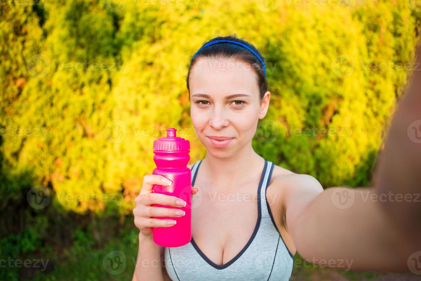 Young woman with bottle of water after running outside. Female fitness model training outside and taking selfie in the park. Healthy wellness fitness lifestyle. photo