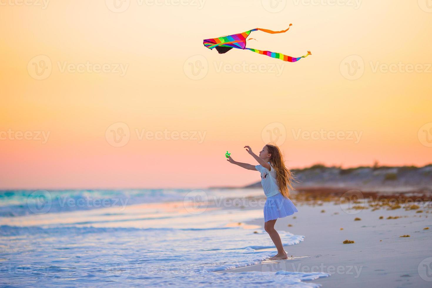 Little girl with flying kite on tropical beach at sunset. Kid play on ocean shore. Child with beach toys. photo