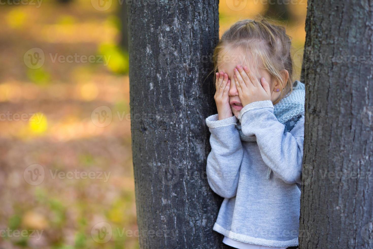 Little girl playing hide and seek near the tree in autumn park photo