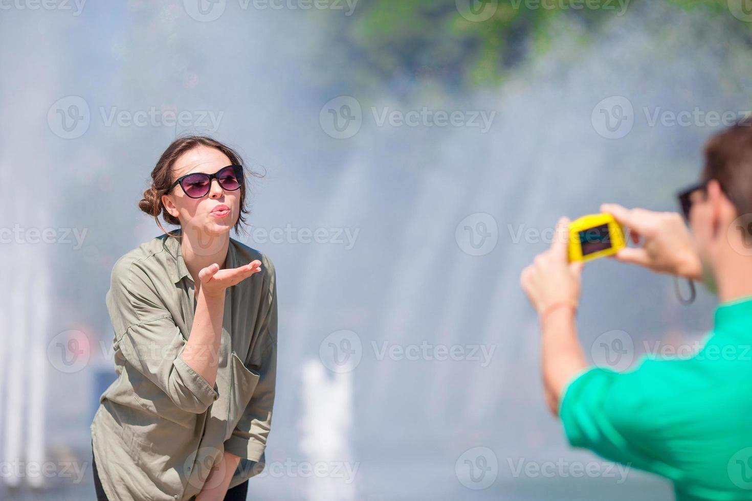 Young man taking a picture of his girlfriend while sitting background the fountain. Young man making photo of woman on the street laughing and having fun in summer.