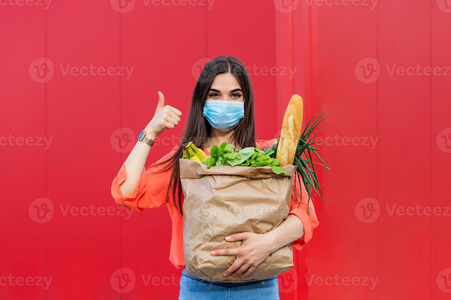 Pretty girl with medical mask holding bag with groceries and looking at camera with thumb up. Woman holding heavy bag with groceries. Groceries shopping during Covid 19, coronavirus pandemic photo