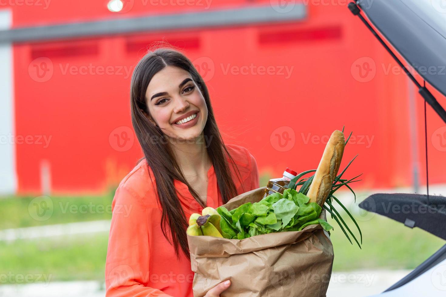 Caucasian brunette going holding paper bags with food products. Young woman putting package with groceries and vegetables into car trunk. Attractive caucasian female shopping in mall or grocery store photo