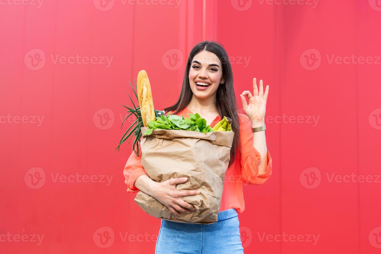 joven mujer hermosa sosteniendo una bolsa de papel de comestibles del supermercado con una cara feliz mostrando el signo de ok de pie y sonriendo con una sonrisa segura foto