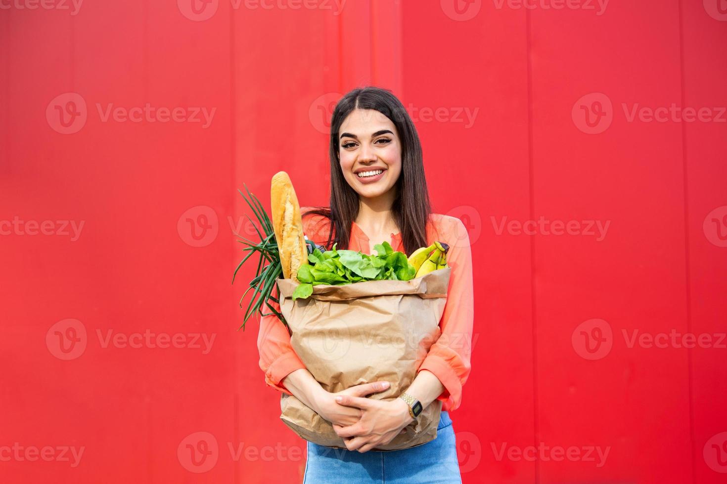 hermosa joven sonriendo sosteniendo una bolsa de papel llena de comestibles. Chica guapa feliz sosteniendo una bolsa con comestibles sobre fondo rojo. foto