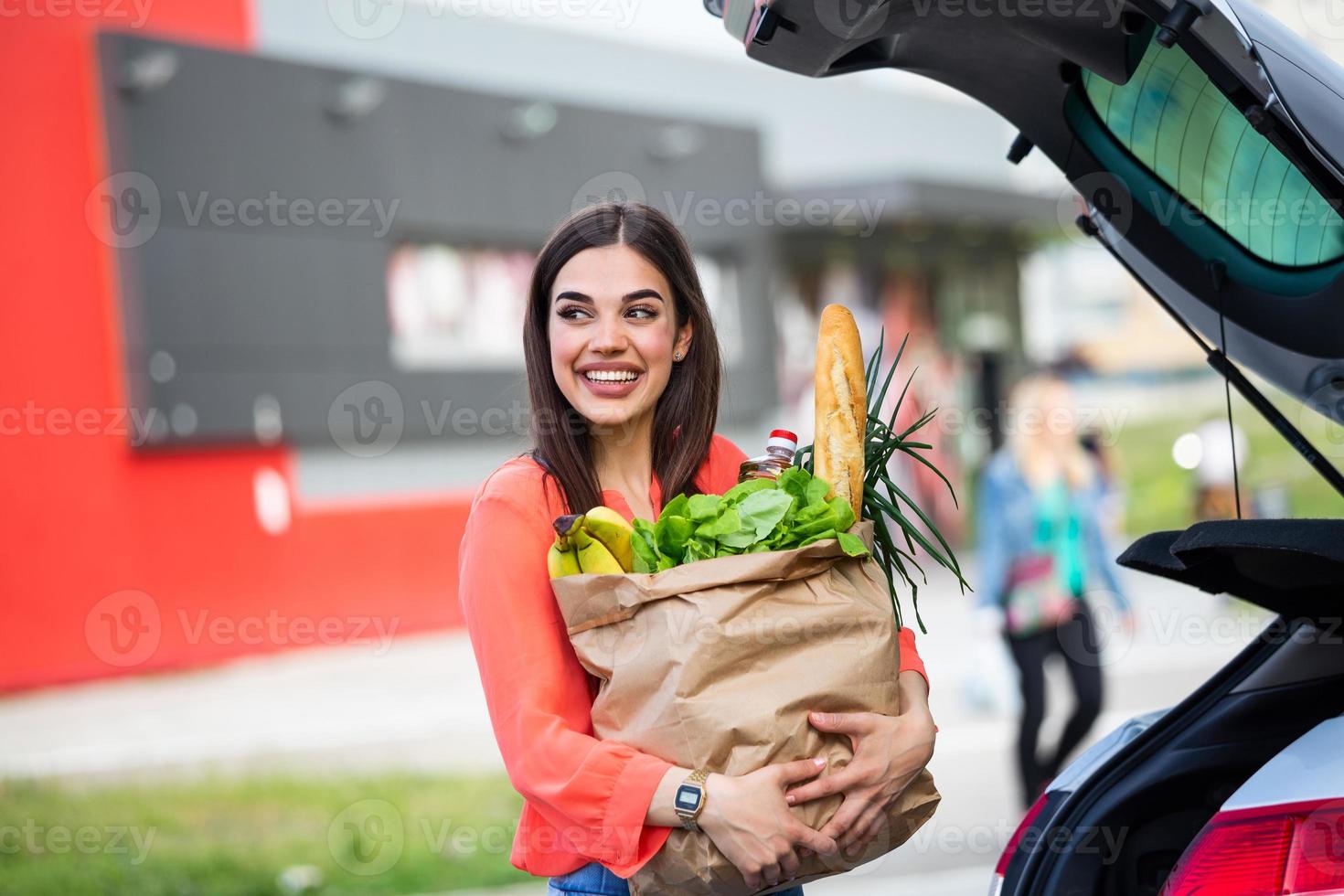 mujer joven montando carrito de compras lleno de comida en el estacionamiento al aire libre. mujer joven en el aparcamiento, cargando las compras en el maletero del coche. Compra realizada con éxito. mujer poniendo bolsas en el coche después de ir de compras foto