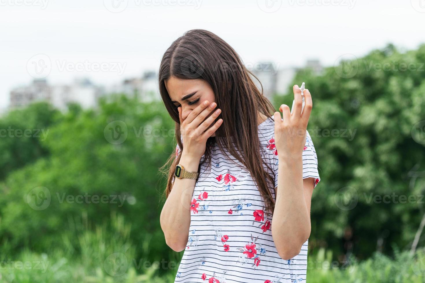 Concept of spring illness. Young female holding napkins and spray for her allergy or fever, cold and running nose in blooming garden. Irritated woman with seasonal allergy photo