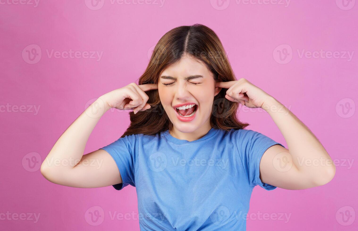 Portrait of angry irritated young woman wearing casual t-shirt covering her ears with hands and shouting while standing isolated over pink background photo