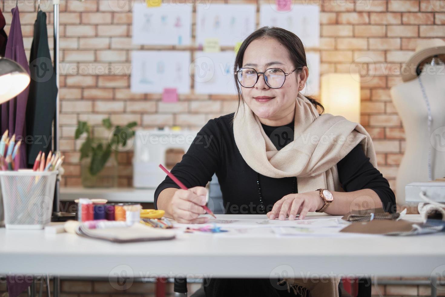 Asian middle-aged female fashion designer working in a studio by idea drawing sketches with colorful thread and sewing for a dress design collection, professional boutique tailor SME entrepreneur. photo