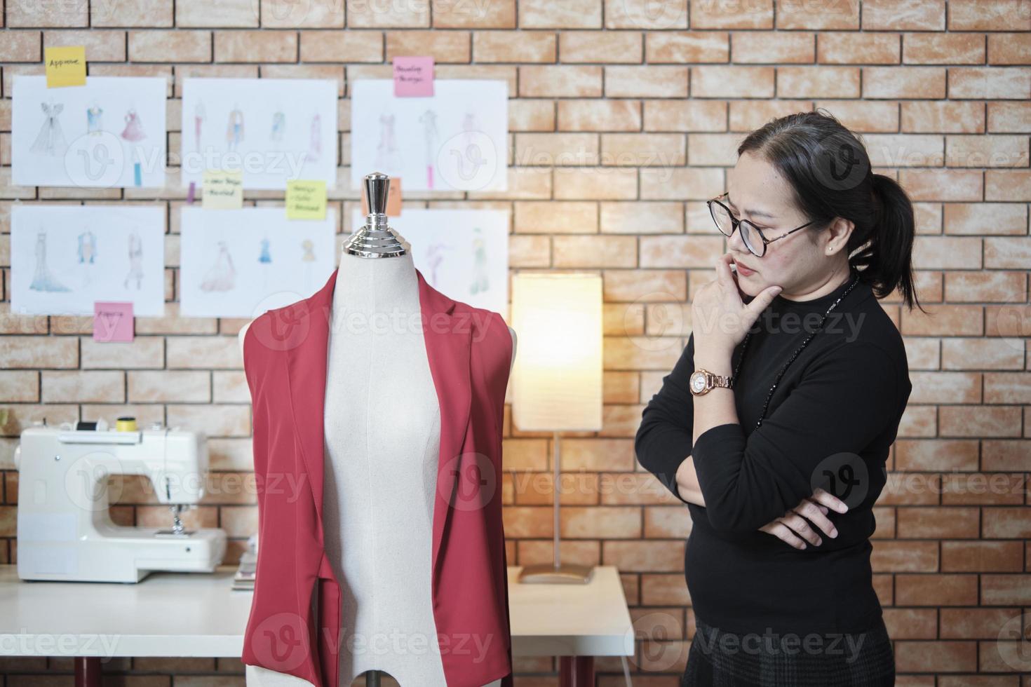 Asian mature female fashion designer stands near dressing puppet, thinking and contemplating for design ideas in studio, works with clothing collection, professional boutique tailor SME entrepreneur. photo
