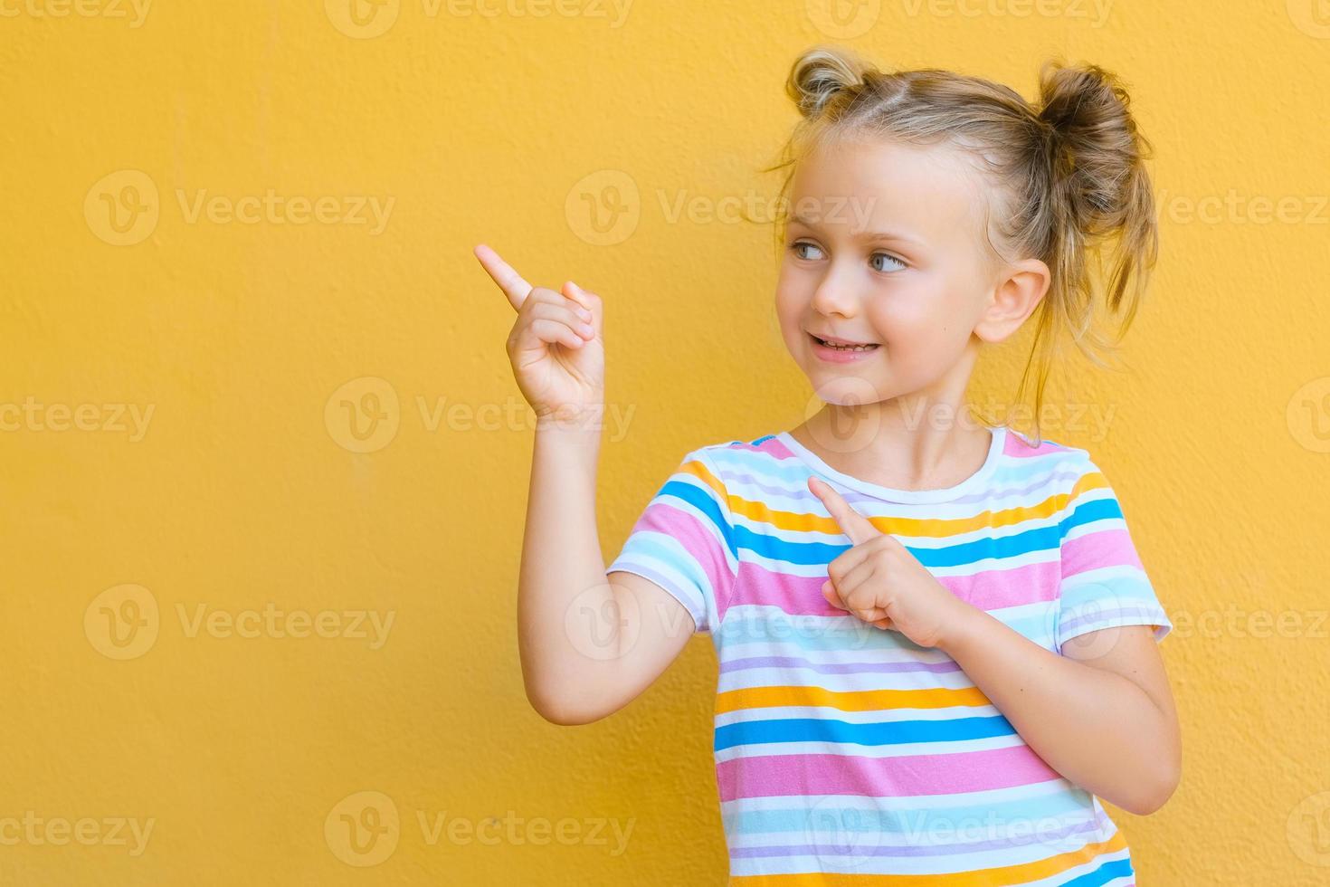 la niña sonriente se siente atraída por la atención que señala con el dedo el espacio de la copia. el concepto de publicidad de productos, bienes y servicios. aislado sobre fondo amarillo foto