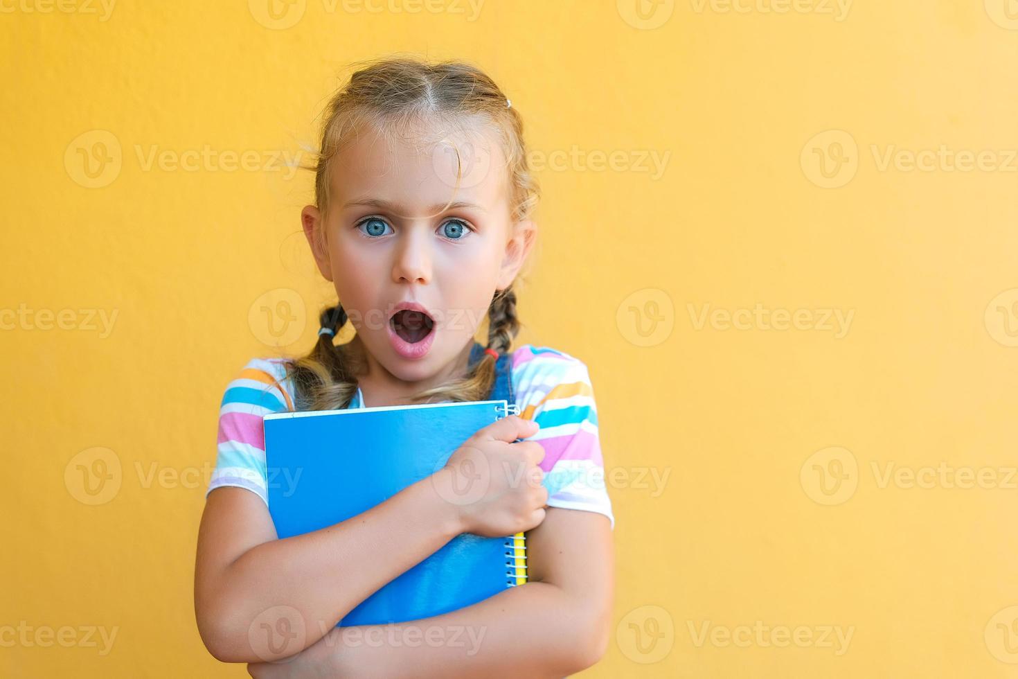 portrait of surprise cute little child school girl in striped T-shirt holds notebook and pen. concept of education,back to school. isolated yellow background. advertising school supplies. mockup photo