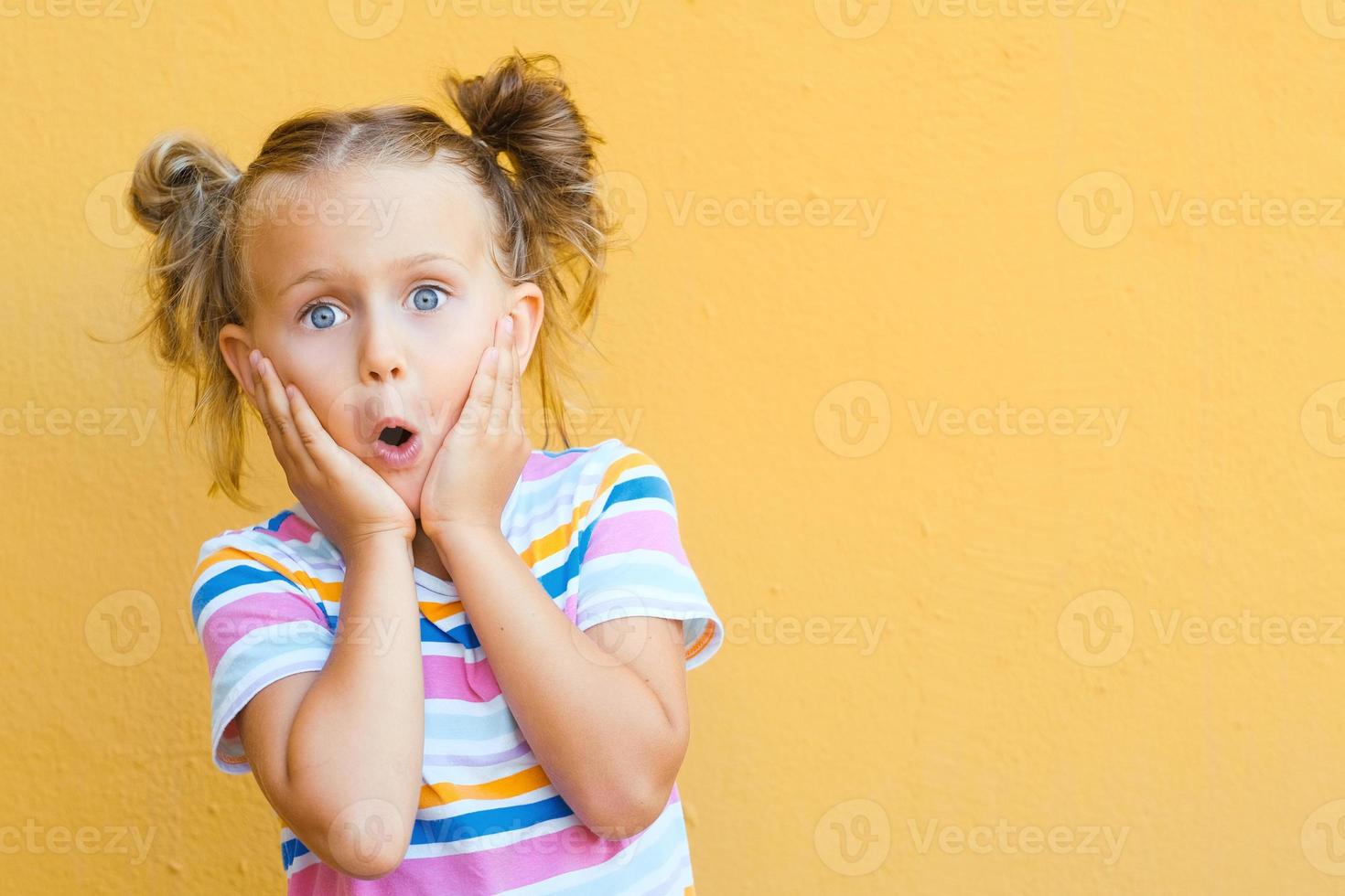 Little child girl in striped colored summer t-shirt surprised expression looks at camera on yellow background, studio portrait.Advertising of children's products and sale photo