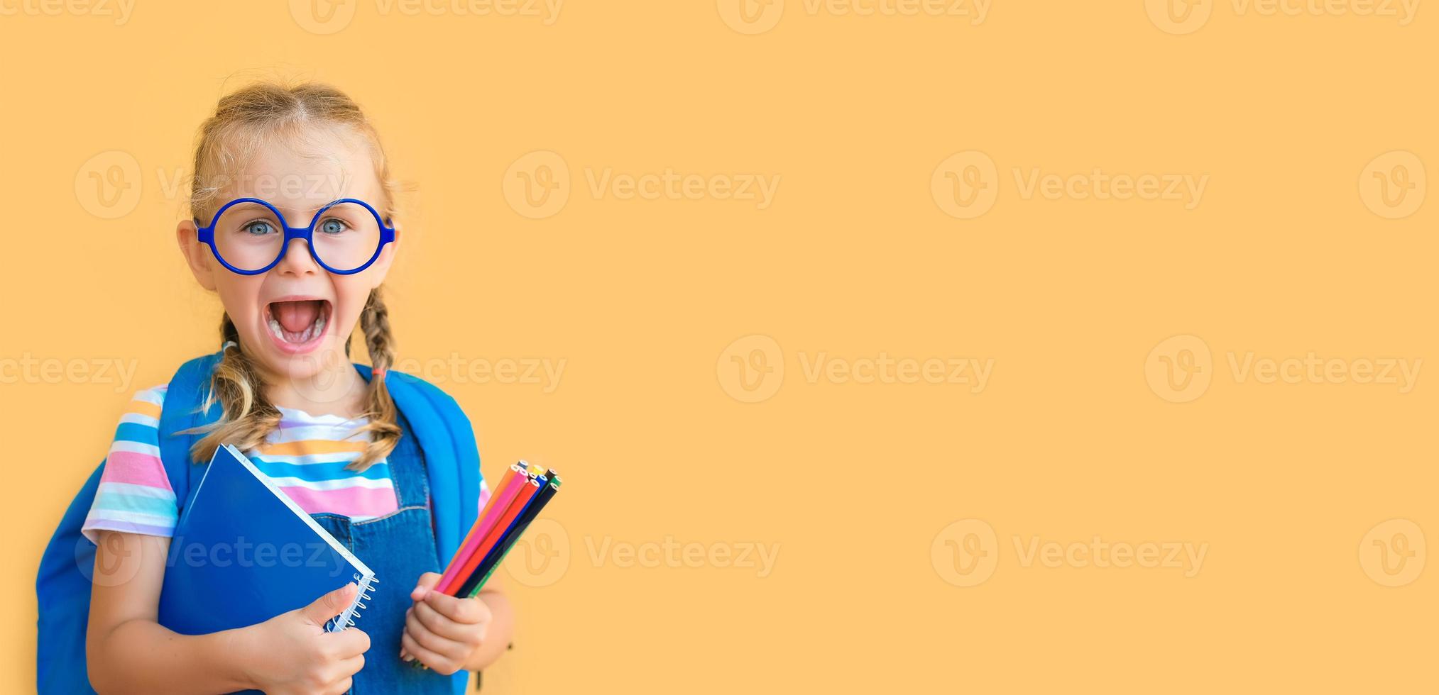 Banner. emotional excited surprise face school child in glasses in school uniform holds colored pencils, notebook, backpack open mouth isolated on yellow background. Back to school sale photo