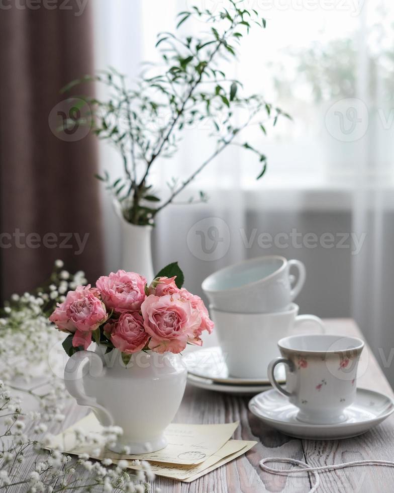 A vase of pink roses and white cups on a table by the window. Spring, summer still life photo