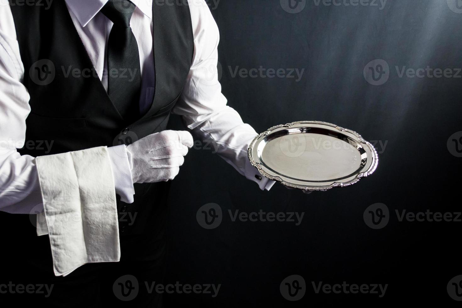 Portrait of Butler or Waiter in Black Vest and White Gloves Holding Serving Tray on Black Background. Concept of Service Industry and Professional Courtesy. photo