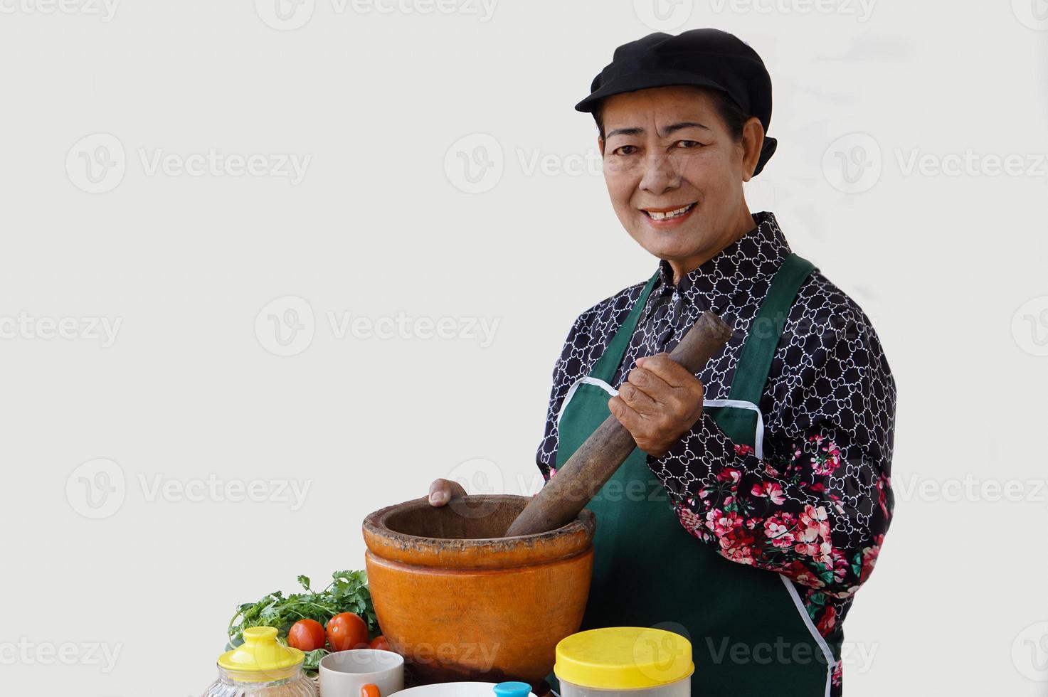 Happy Asian senior woman is cooking, wear chef cap and apron, holds pestle, mortar and plate of chillies. Concept, Cooking for family. Thai kitchen lifestyle. Elderly activity. photo